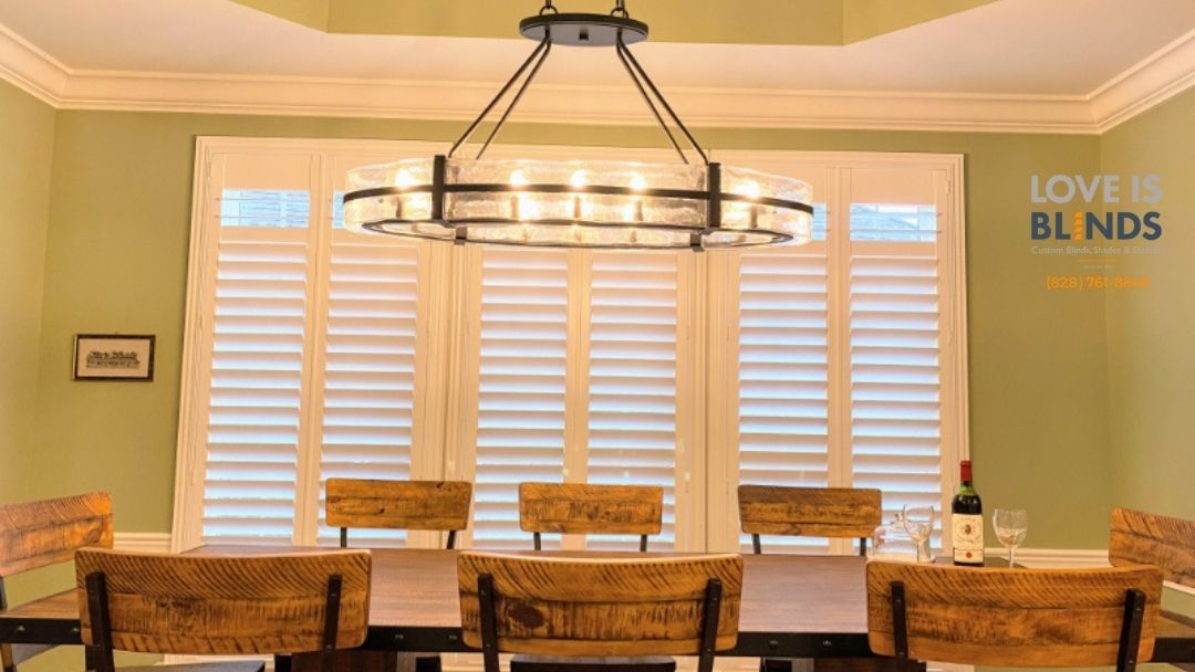 Dining room with wooden table, chairs, and shutters; a chandelier hangs above.