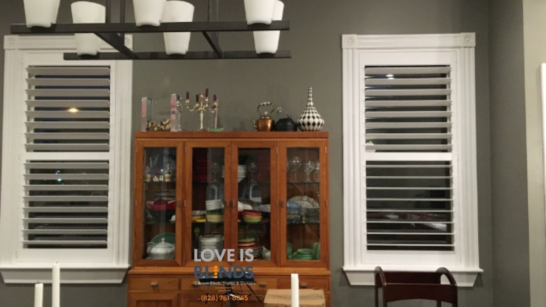 Dining room with wooden hutch, window shutters, and chandelier against a gray wall.