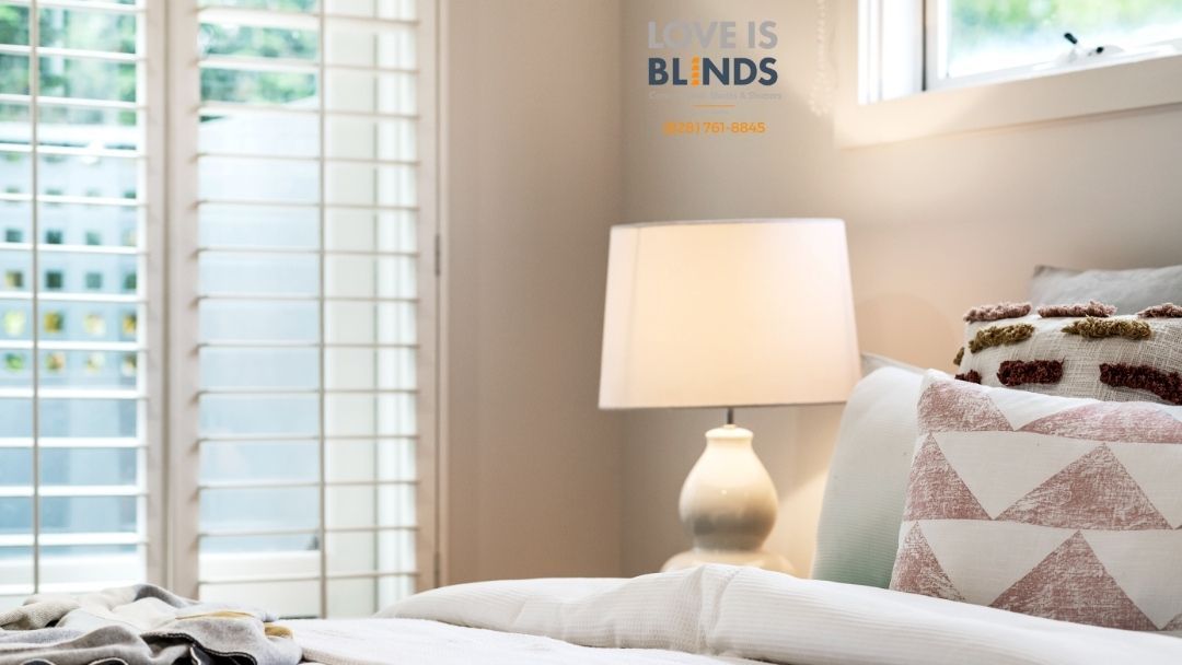 Bedroom with white shutters, lamp on nightstand, pillows on bed.