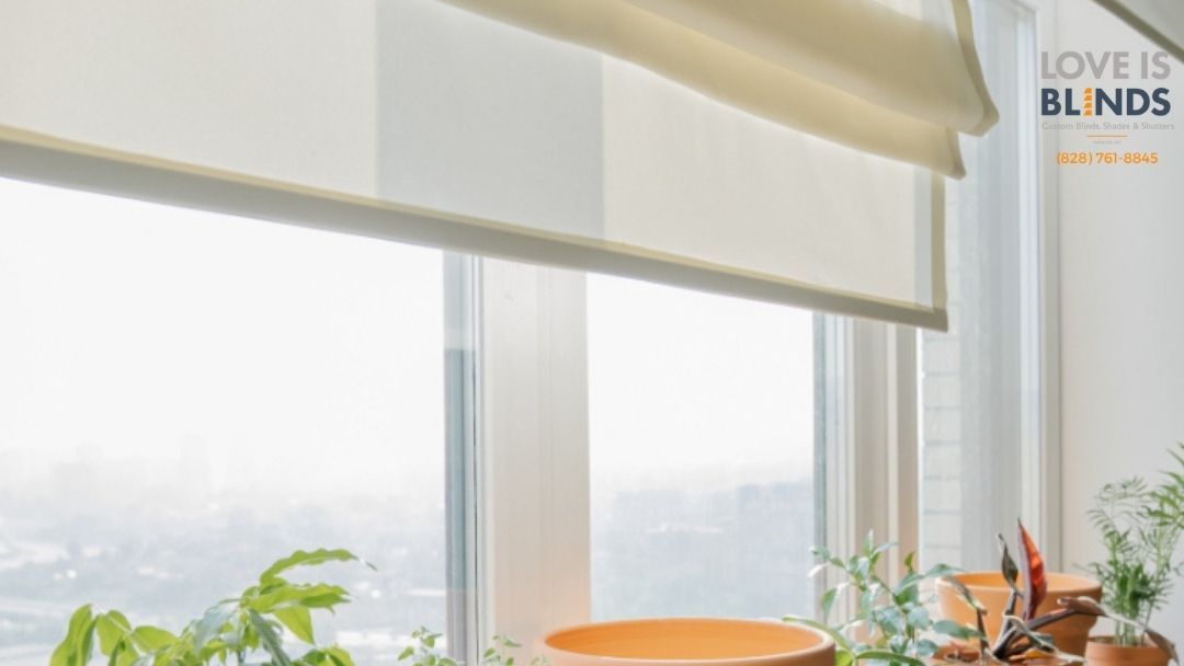 Cream-colored roller blinds partially covering a window, overlooking a bright outdoor scene with plants on the windowsill.