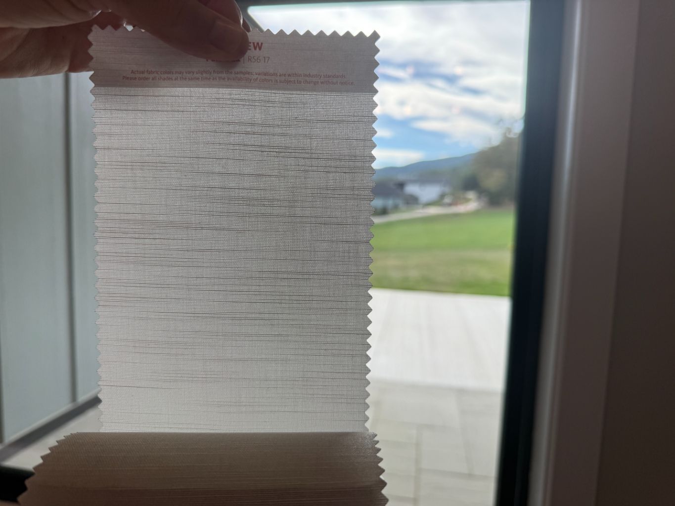 Hand holding a white textured fabric swatch for roller shades up to a bright window, with a yard visible outside