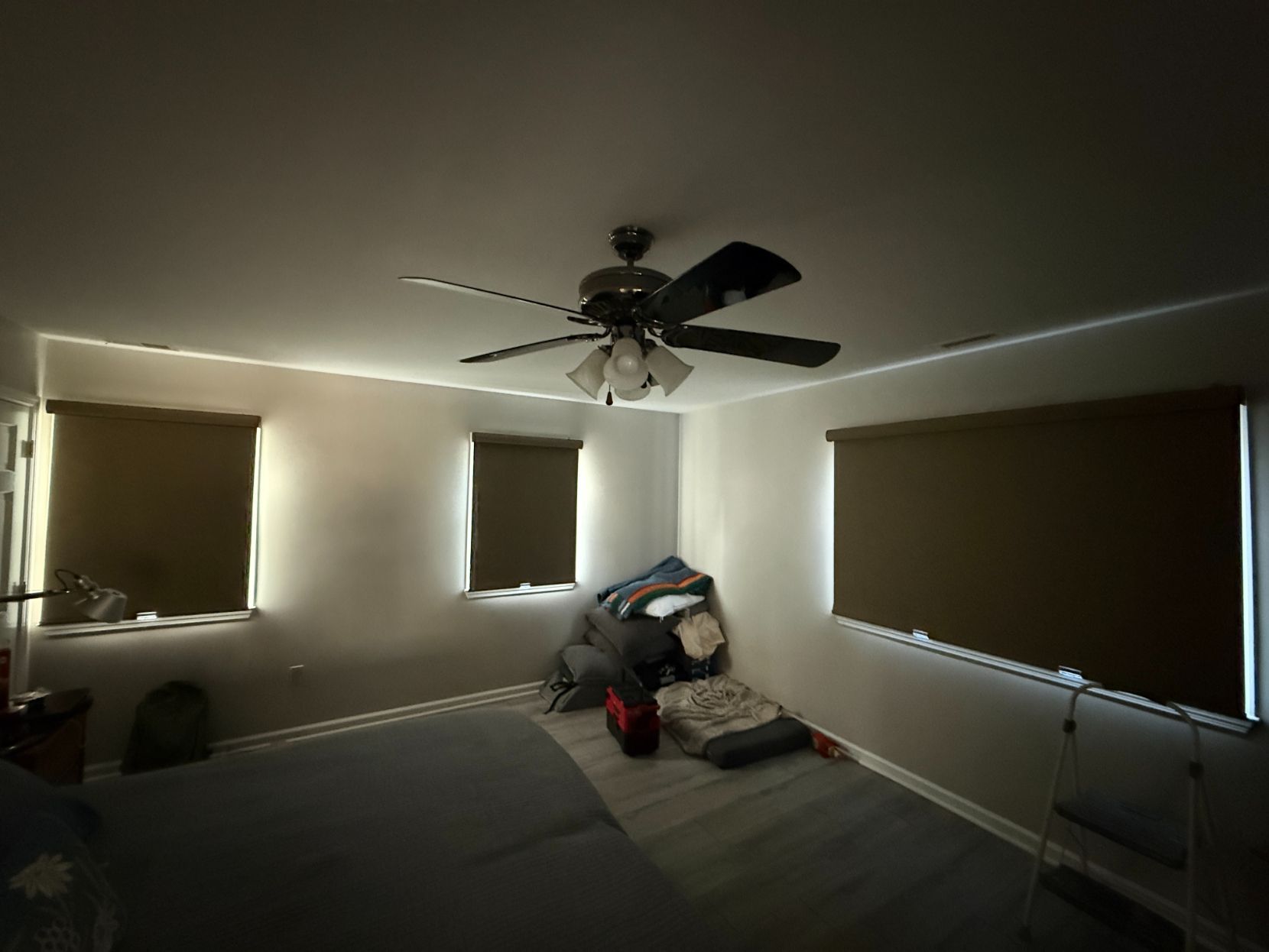 Dim bedroom with blackout shades closed, ceiling fan, and a pile of clothes on the bed.