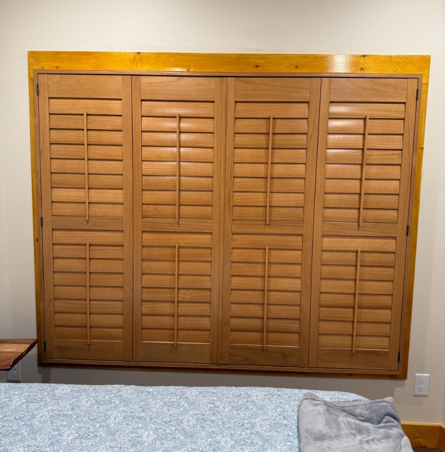 Four light-wood plantation shutters installed on a window above a bed with a blue patterned comforter.