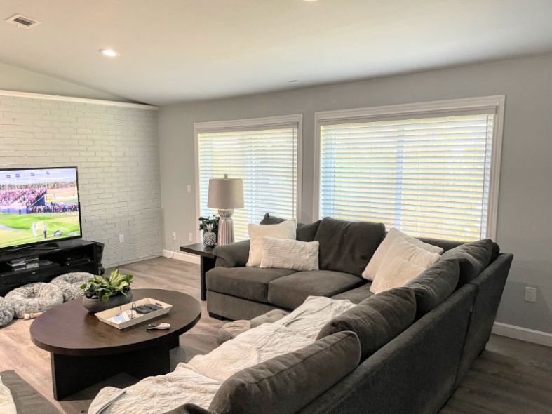 Living room with gray sectional sofa, TV, coffee table, and large windows with blinds