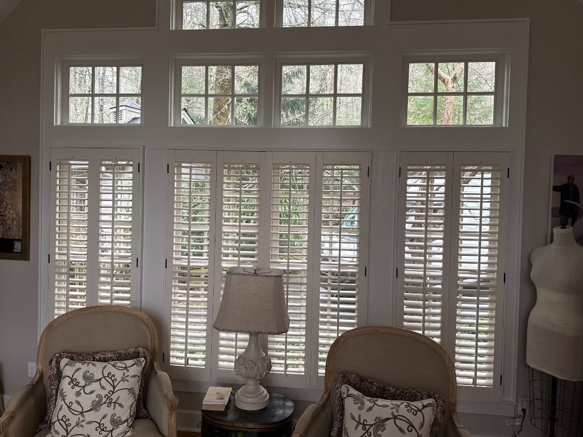 Bright sitting room with tall white shutters, two wicker chairs, and a table lamp centered between them