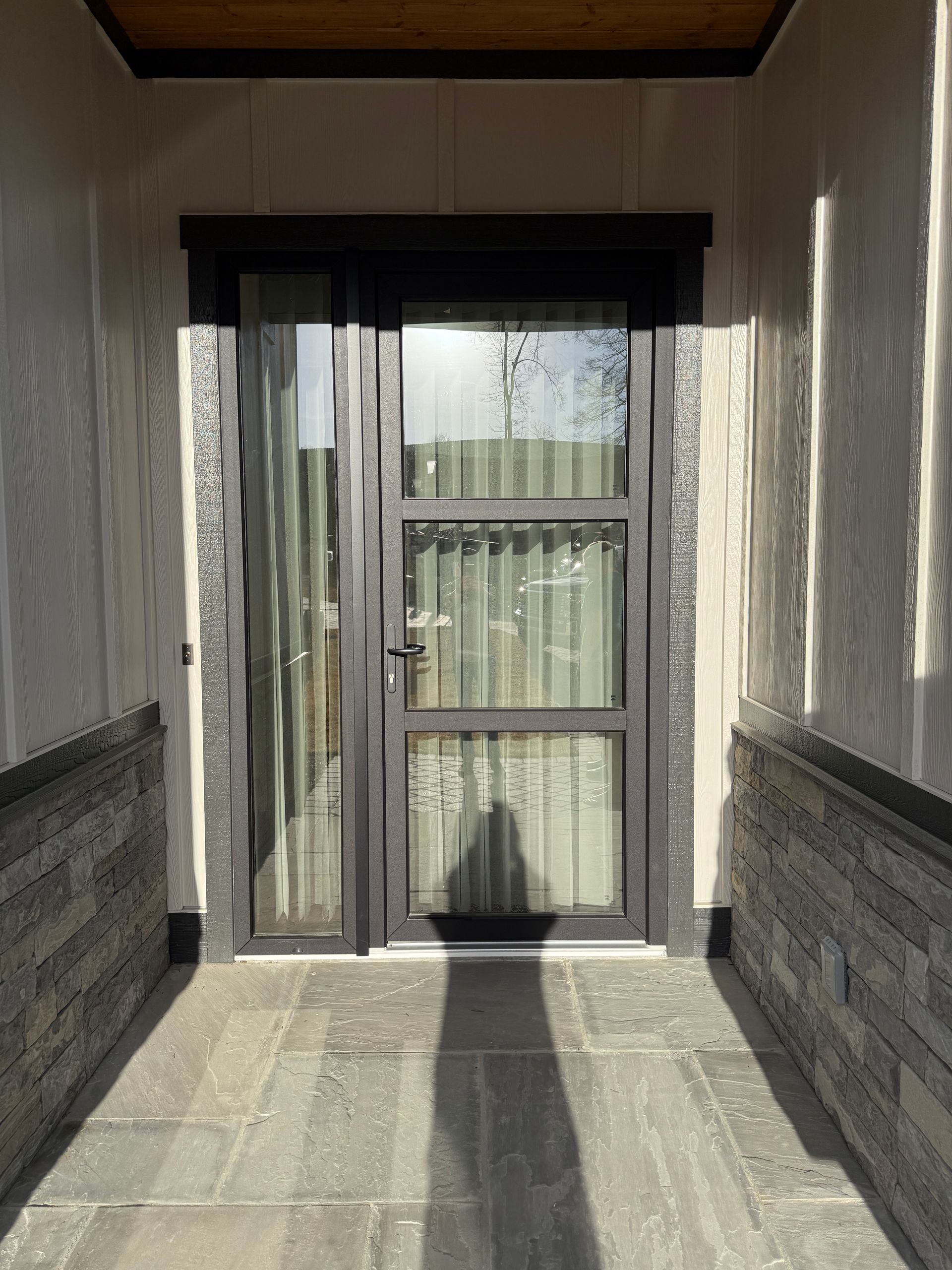 A dark-framed entry door with side glass panel, flanked by stone walls and tile flooring in a covered entryway.