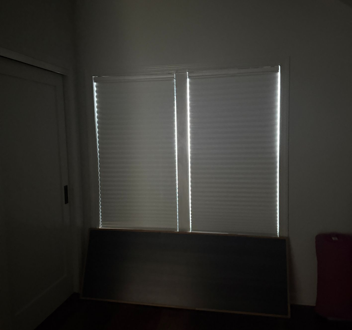 Two windows with white honeycomb shades on a wall above a dark, horizontal piece of furniture in a dimly lit room.