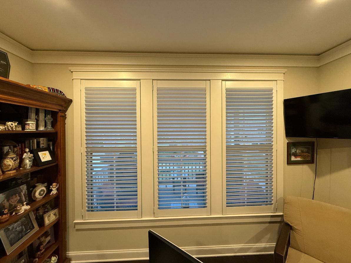 Living room with three windows, wall-mounted TV, beige couch, and plantation shutters.