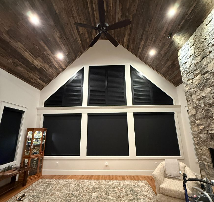 A tall living room with dark wood-plank vaulted ceilings, stone fireplace, and windows covered by black roller shades.
