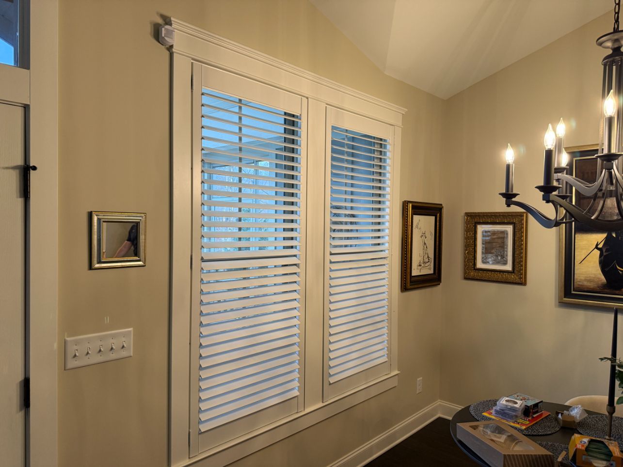 Small beige hallway with white window blinds, framed wall art, and a dark chandelier on the right