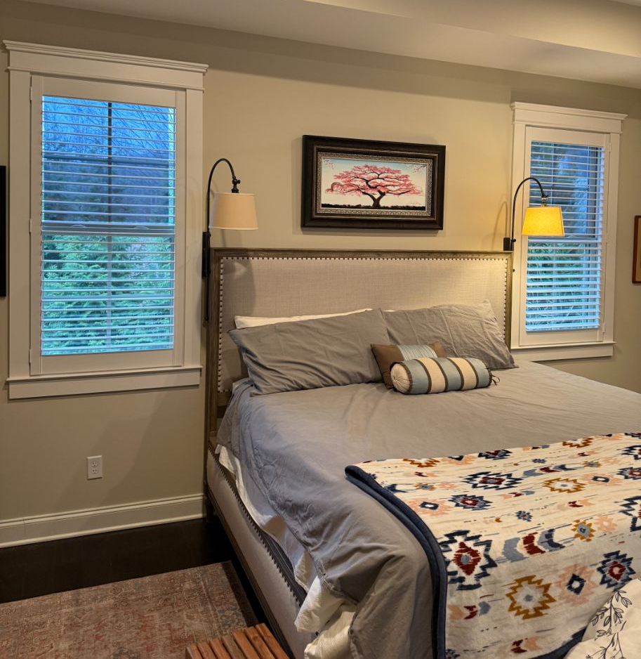 Cozy bedroom with a bed, patterned quilt, gray pillows, wall art, and windows with blinds and lamps.