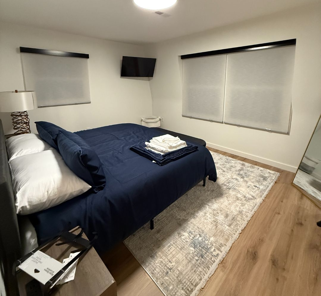 A modern bedroom with a navy blue bed, white walls, light wood floors, a patterned rug, and windows with roller shades.