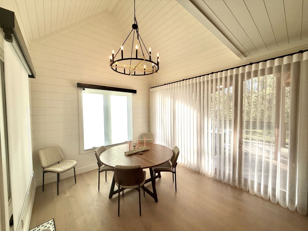 Bright dining room with round table, chairs, chandelier, and sheer curtains beside large windows.