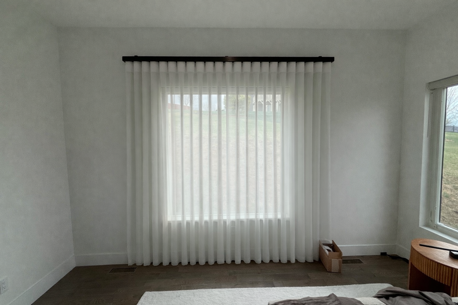 White vertical blinds on a dark rod covering a large window in a room with white walls and dark carpet.
