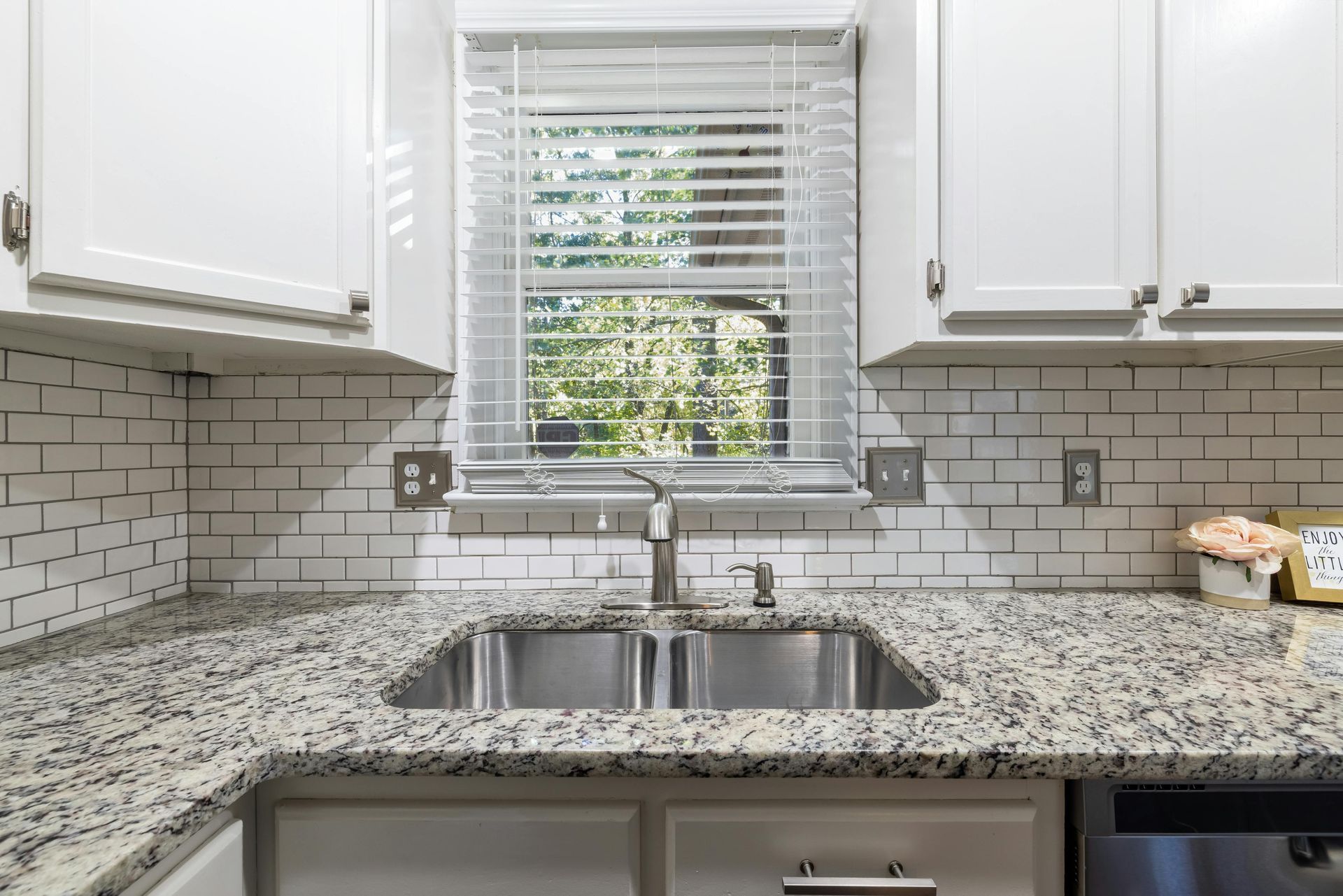 Kitchen sink with granite countertop and white cabinets, window with blinds.