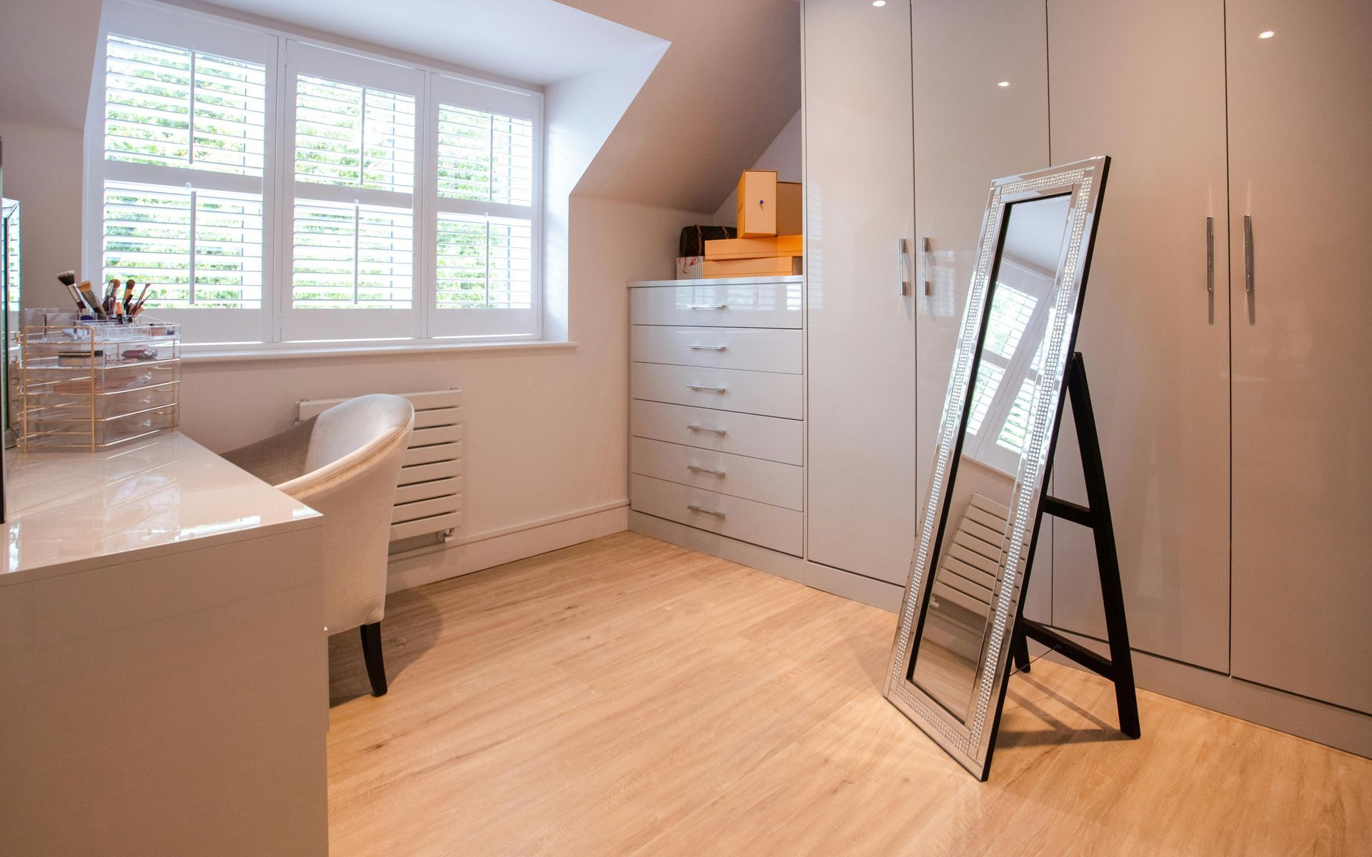 Dressing room with white furniture, large mirror, and window. Light wood floor and neutral walls.