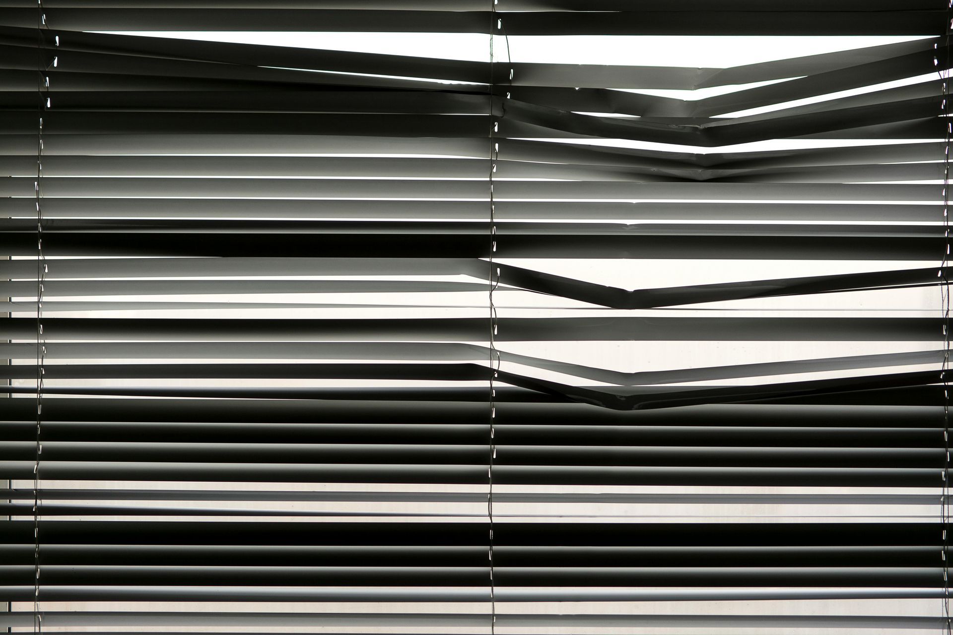 Damaged, closed horizontal blinds with bent slats, letting in some light.