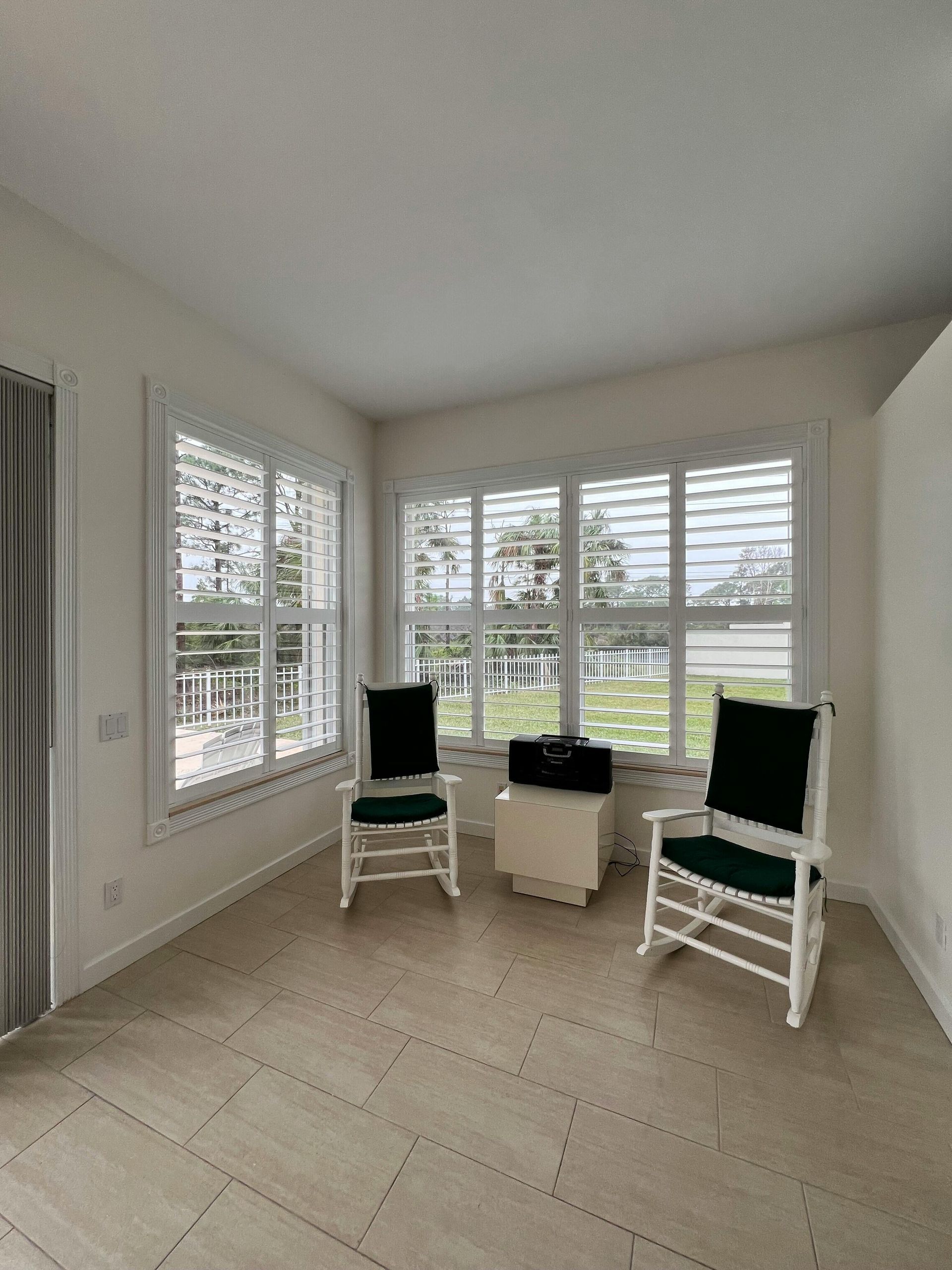 Sunroom with white shutters, two rocking chairs, beige floor, and a small cabinet.