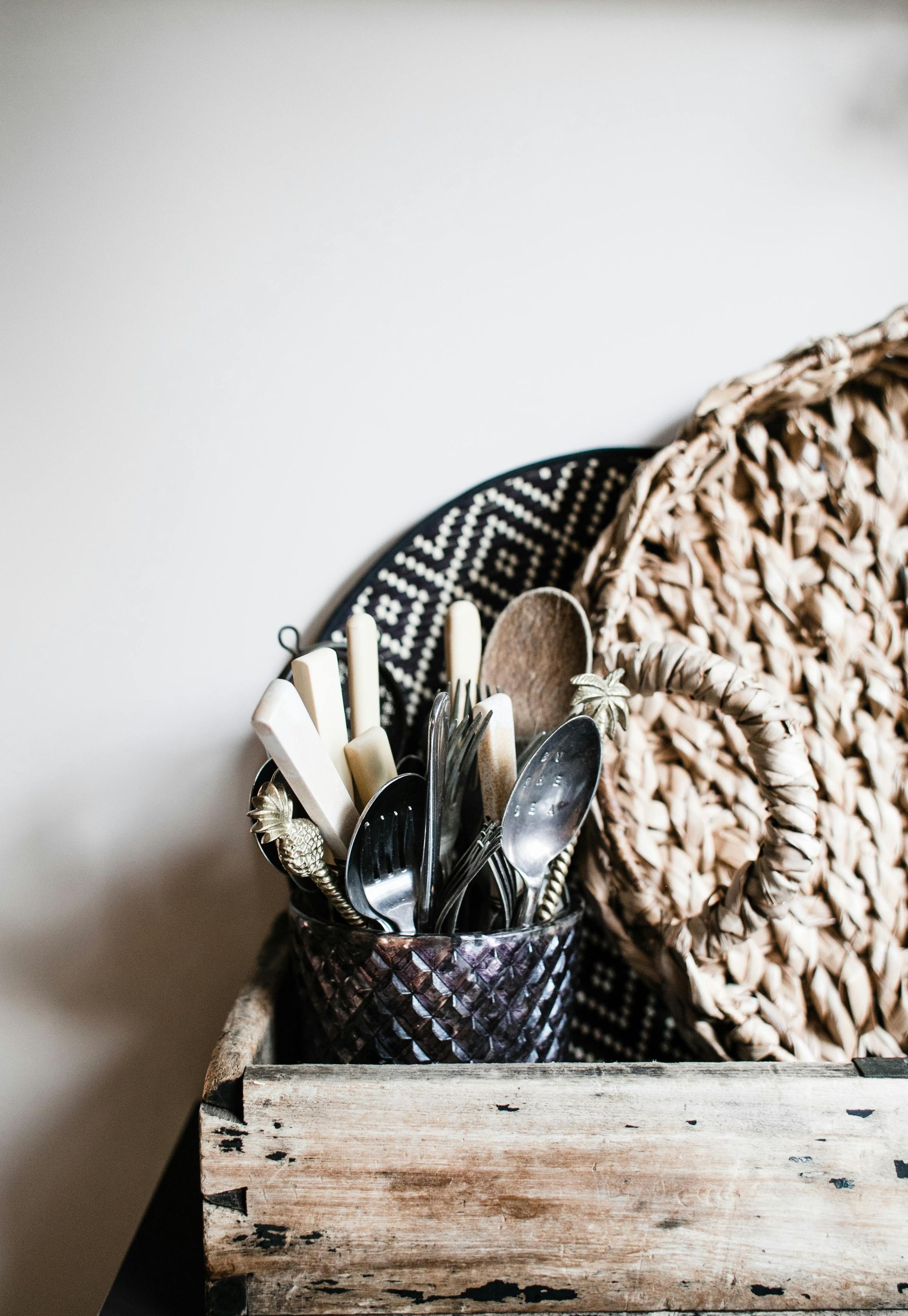 Woven basket holding utensils: spoons, butter knives, and a wooden spoon, on a rustic wooden shelf.