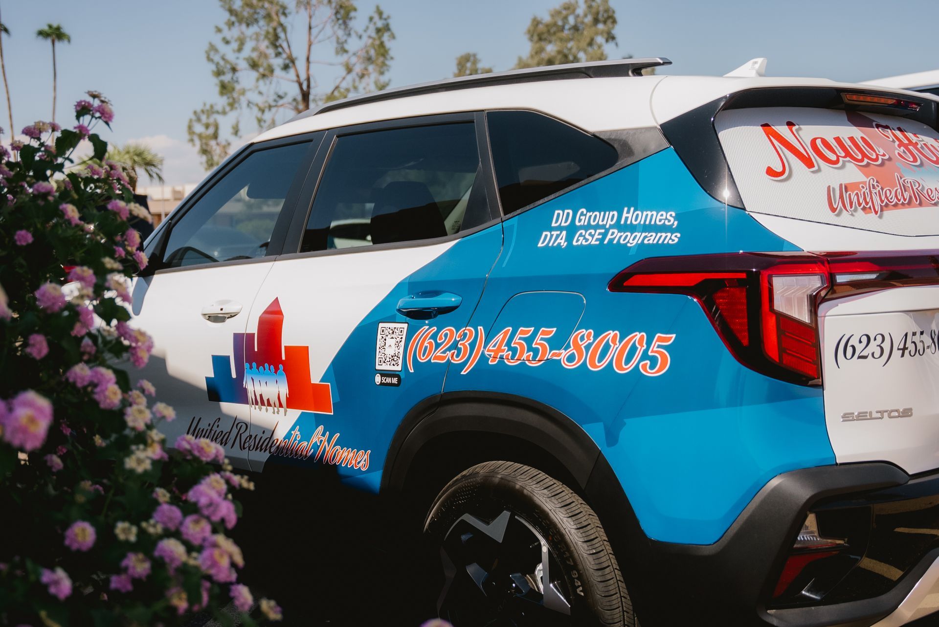 A blue and white URH suv is parked in front of a bush with purple flowers.