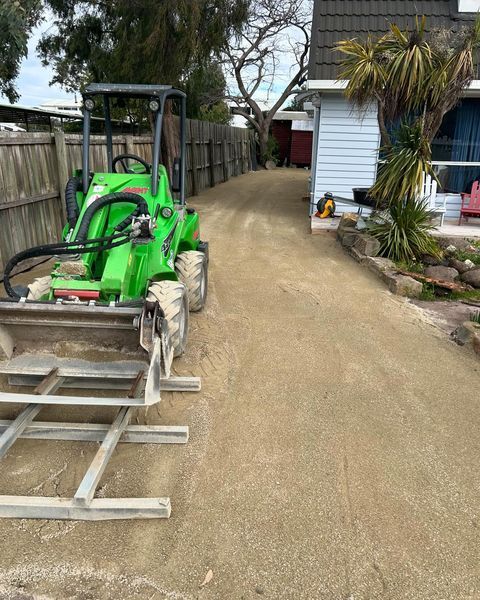 A green tractor is driving down a dirt road in front of a house.