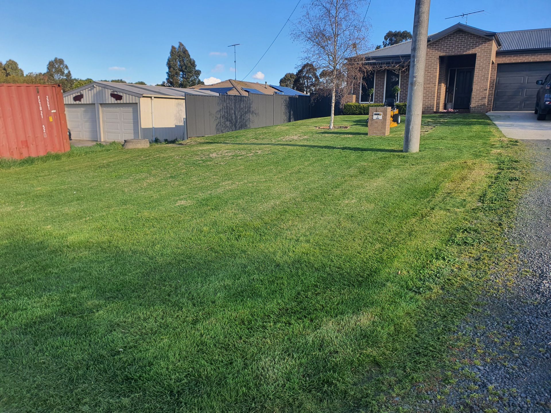 Grassy front yard with a house on the right, a red container on the left, and a cloudy sky — JKR Garden Maintenance Services in Ballan, VIC