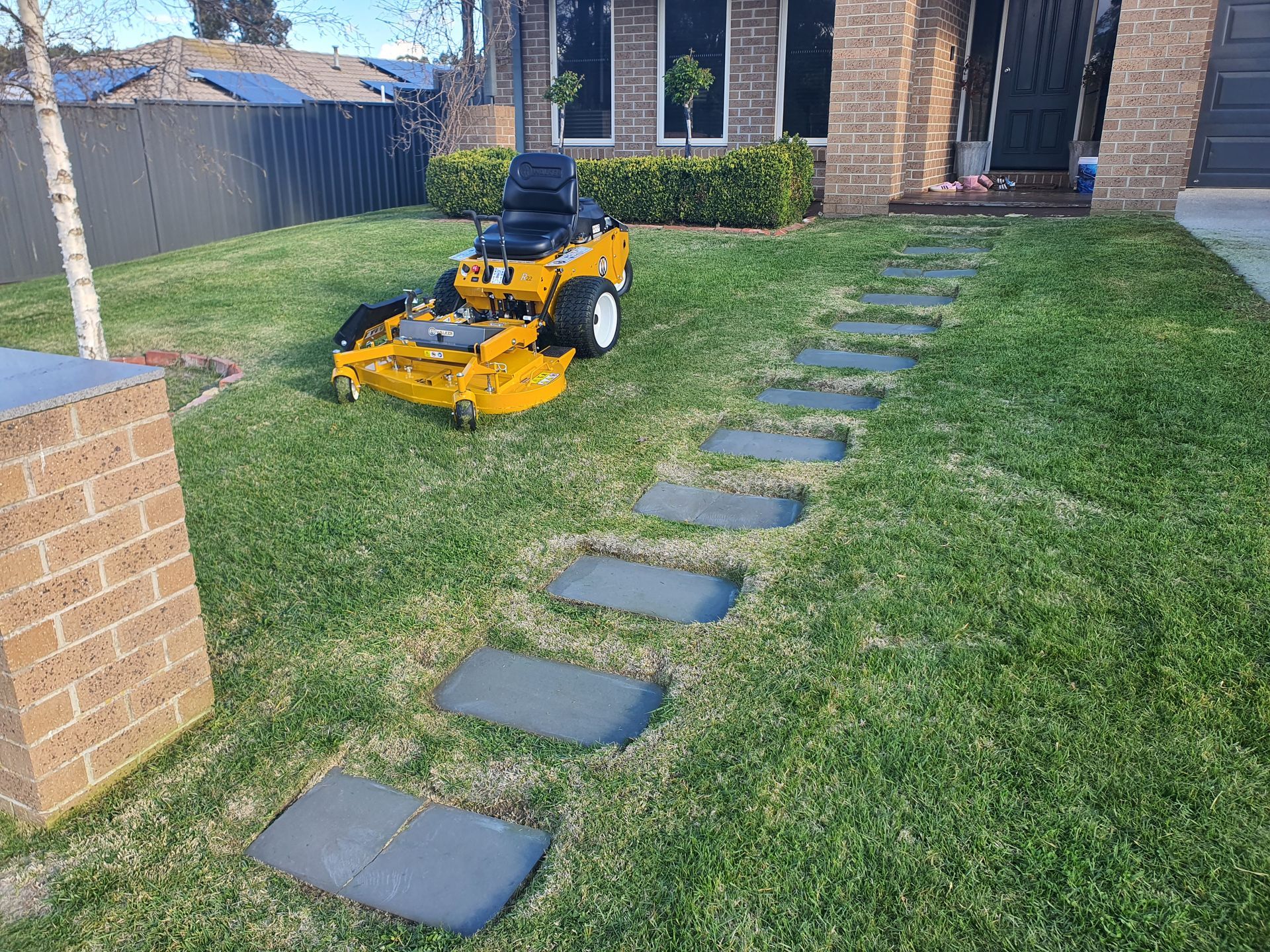 Yellow riding lawn mower on a green lawn, next to a stone pathway in front of a brick house — JKR Garden Maintenance Services in Ballan, VIC