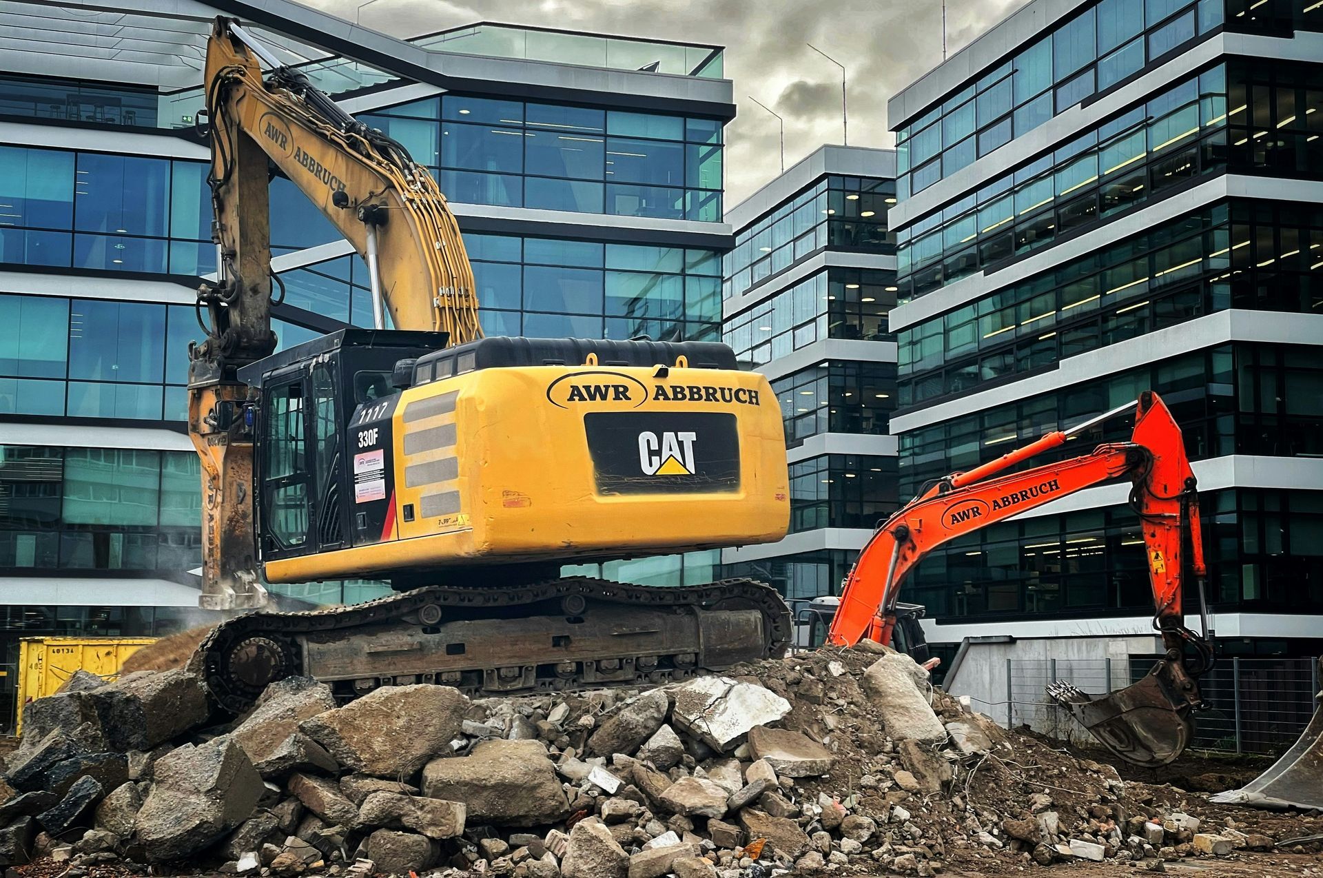 Yellow CAT and orange construction excavators atop a pile of rubble in front of modern glass office buildings.
