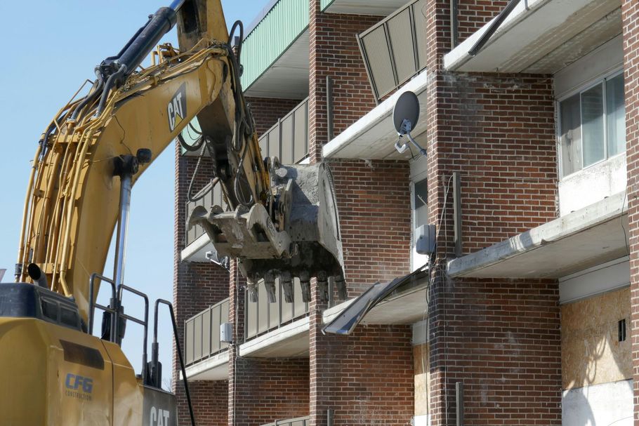 A yellow Caterpillar excavator arm with a metal bucket positioned to demolish a brick apartment building.