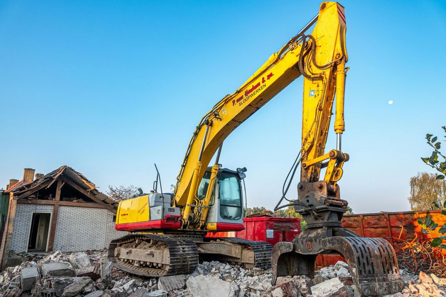 A yellow tracked excavator with a demolition grapple attachment sits atop a pile of rubble near a ruined building.