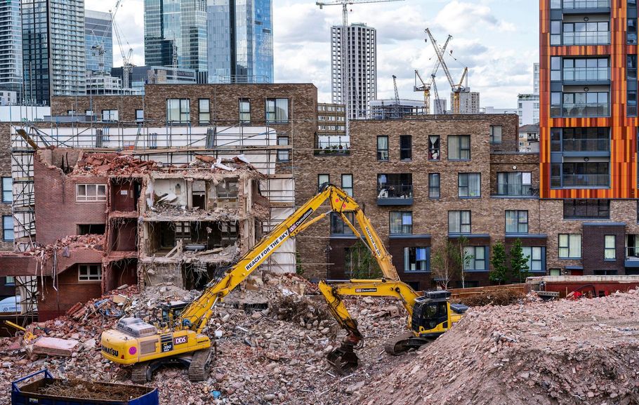 Yellow excavators tear down a brick building amidst a construction site with modern high-rise apartments in the background.