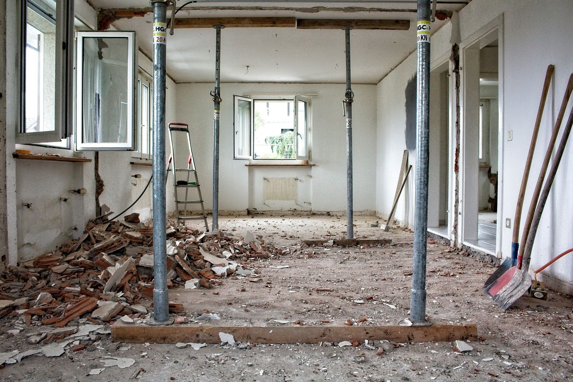 An interior room under renovation with debris on the floor, supported by metal shoring posts and wooden beams.