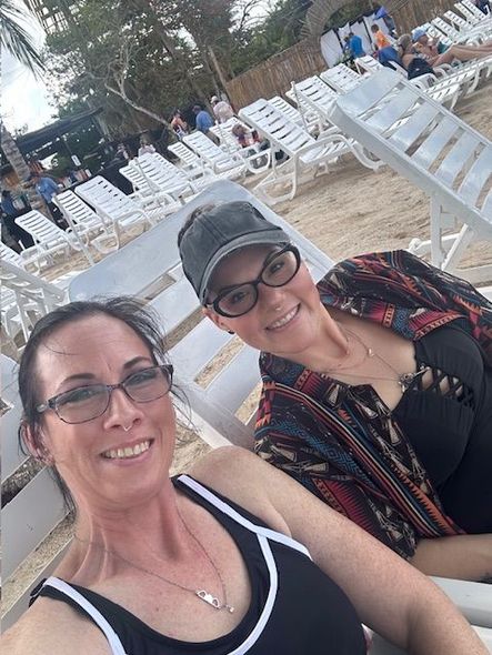 Two people smile, seated on beach chairs; sand, ocean, and other beachgoers in background.