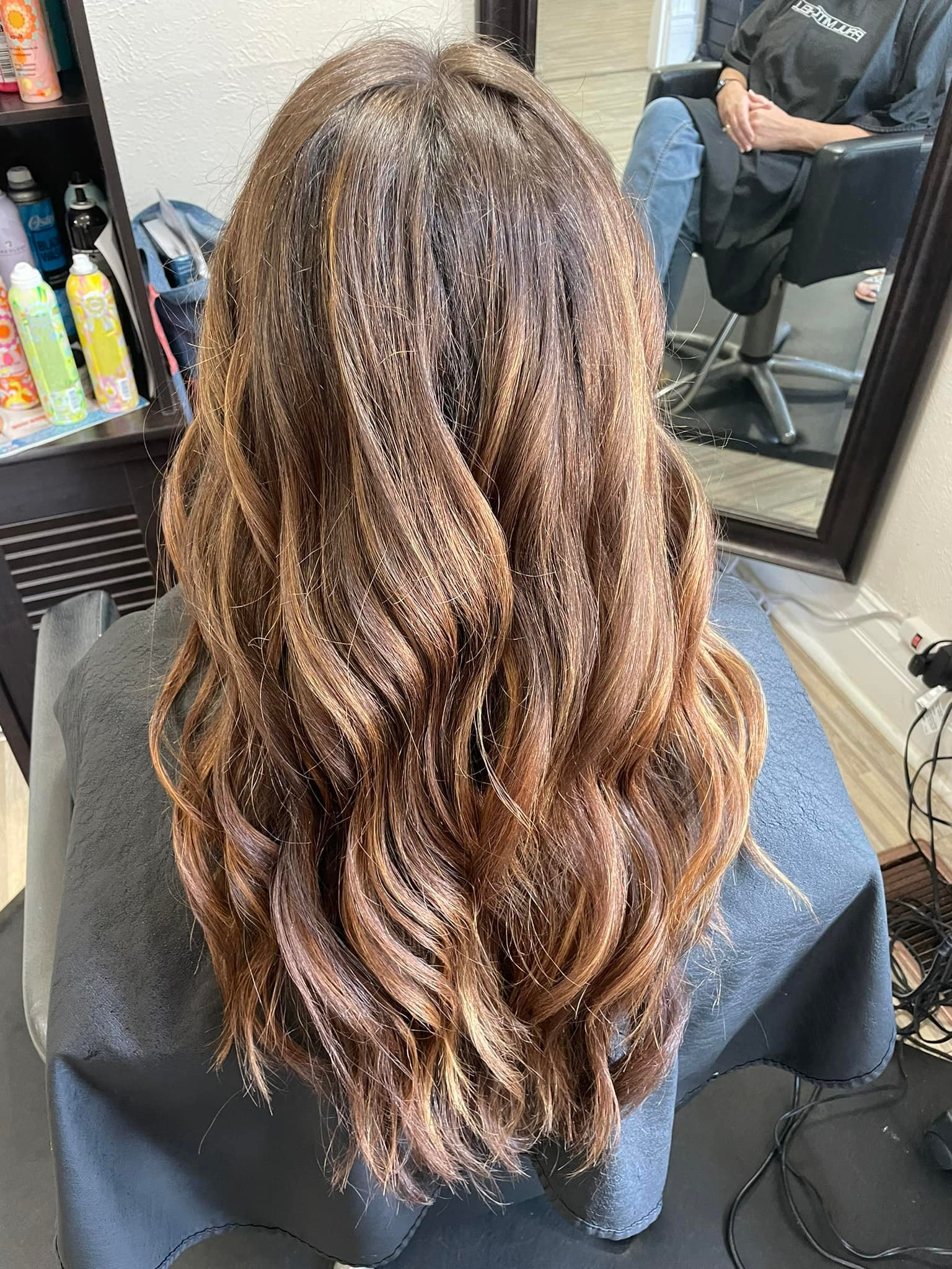 Woman's long, wavy hair with varying shades of brown. She is seated in a salon, draped with a styling cape.