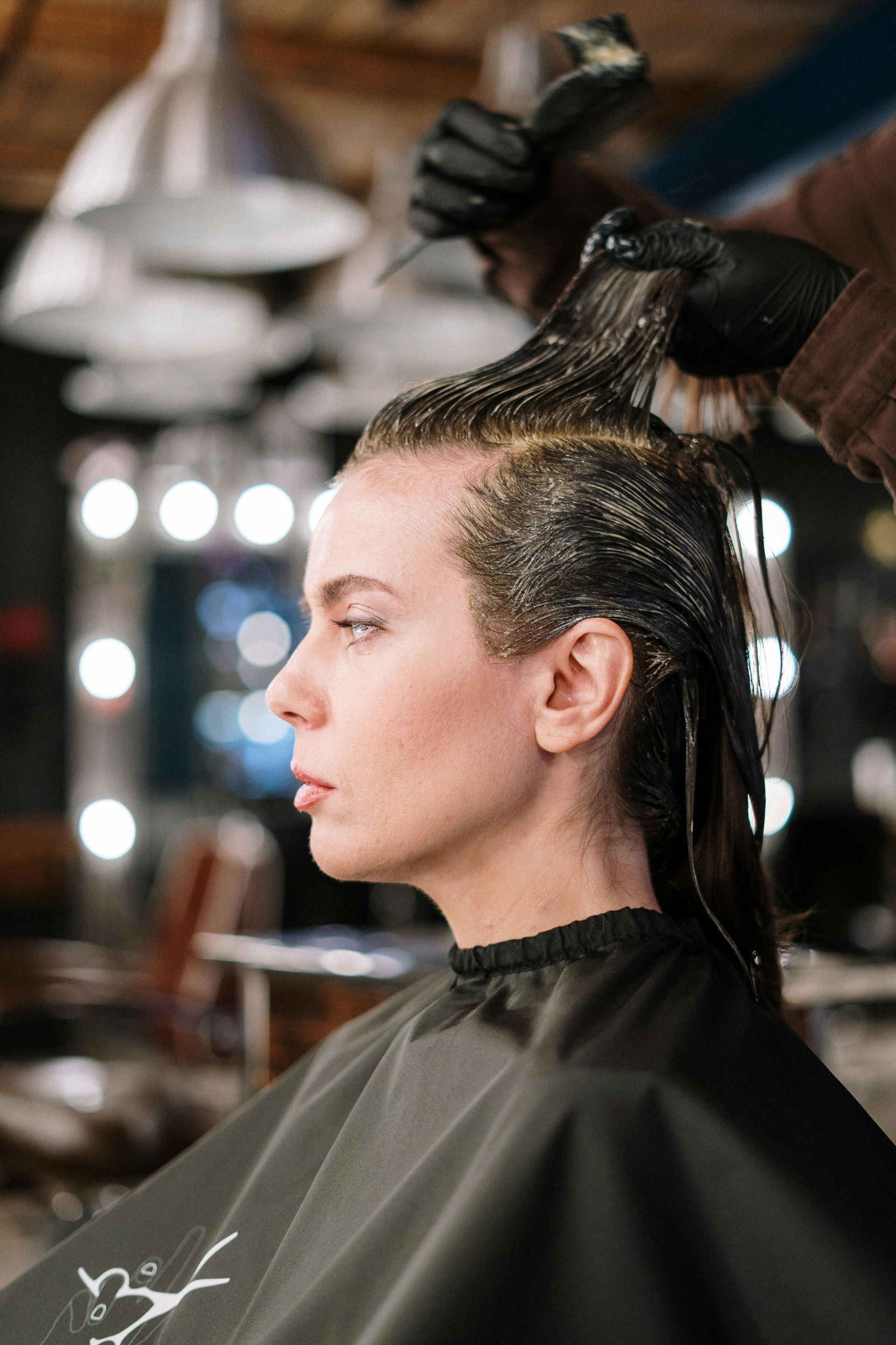 Person having hair dye applied by gloved hand in a salon. Black cape, lights in background.
