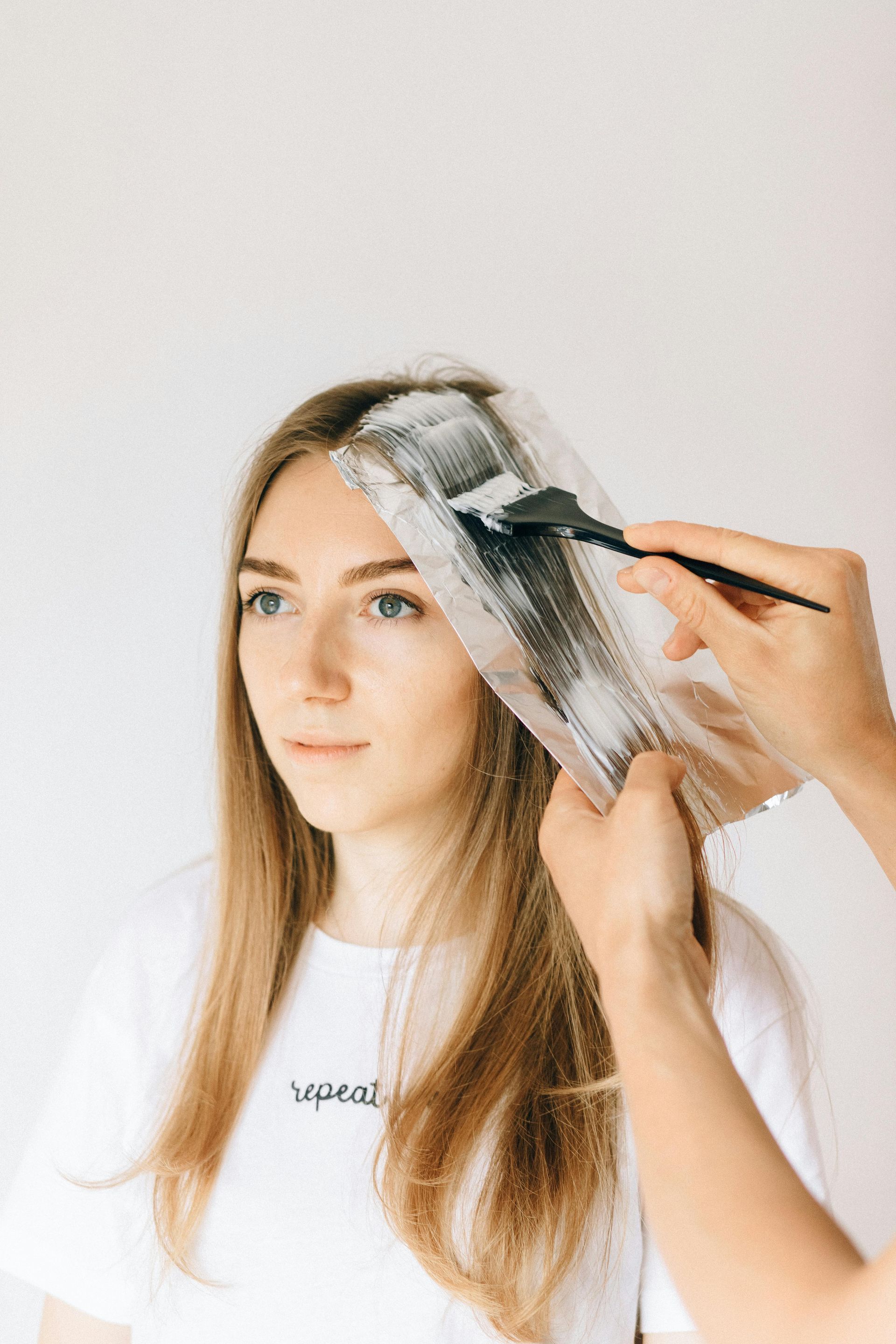Person getting hair highlighted; white cream applied to strands with brush on foil, against white backdrop.