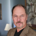 A man with a beard is wearing a brown jacket and black shirt