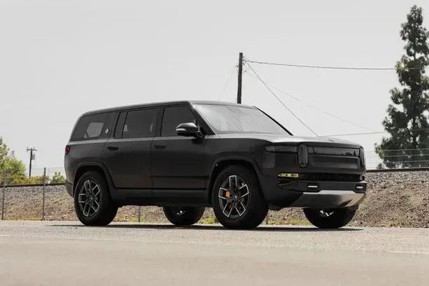 Black Rivian SUV parked on a paved road, facing right, with trees and utility poles in the background.