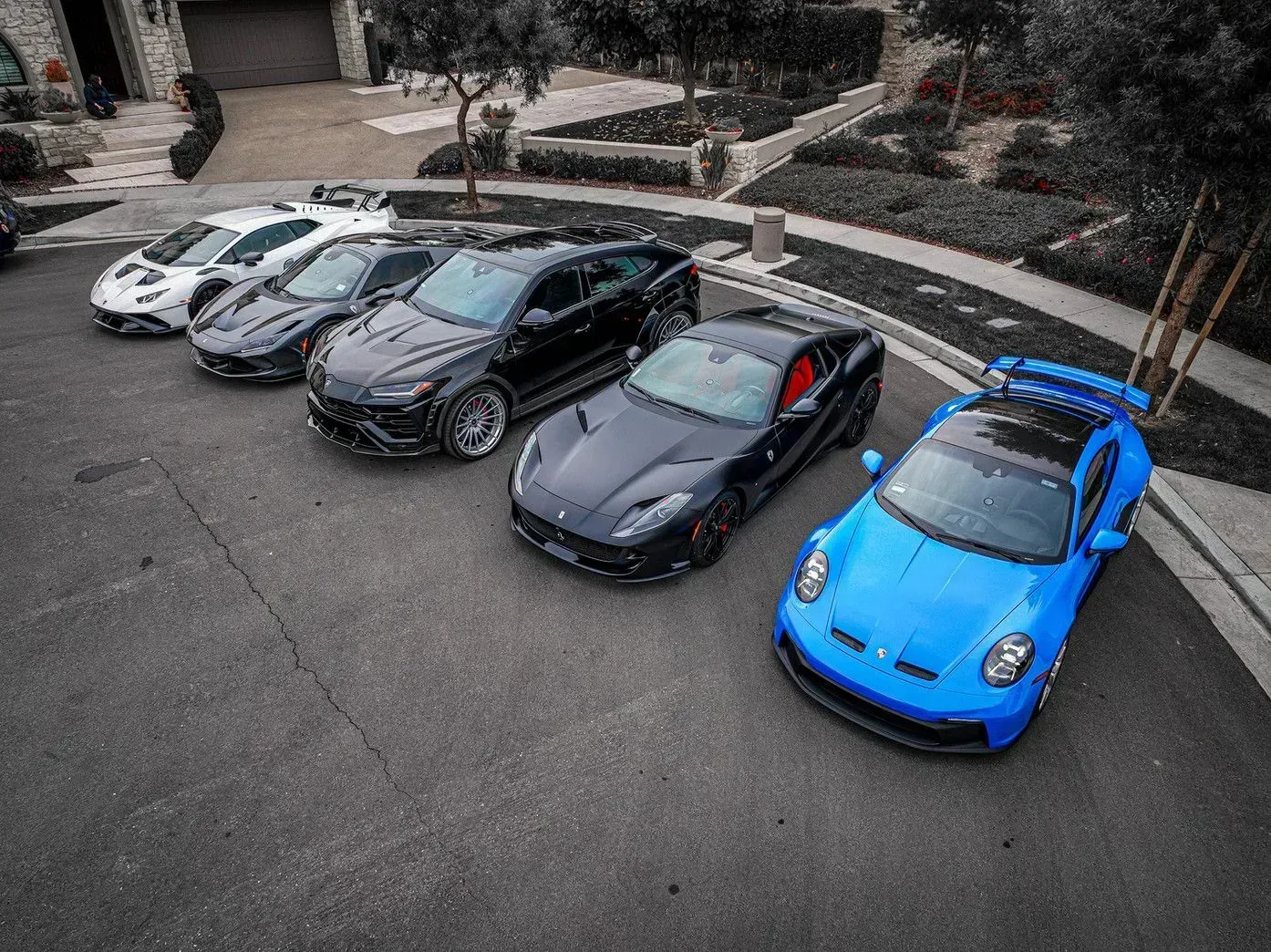 Six luxury cars parked on a driveway; various colors including blue, black, and white.