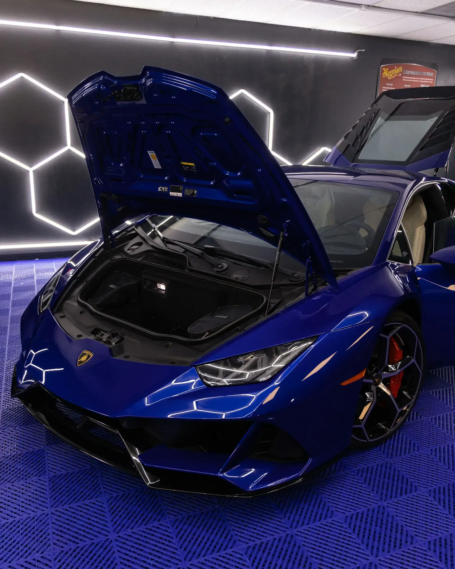 Blue Lamborghini with hood and door open, parked on blue tiled floor under hexagonal lights.