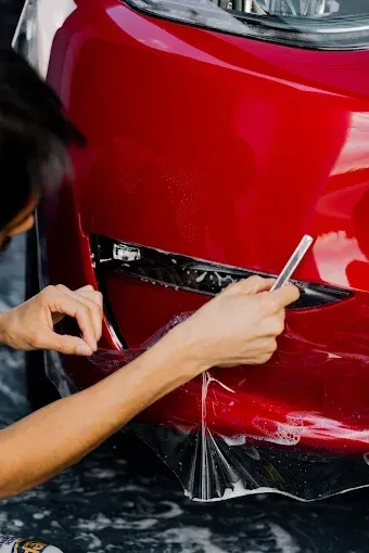 People applying clear protective film to a black car's hood in a garage.