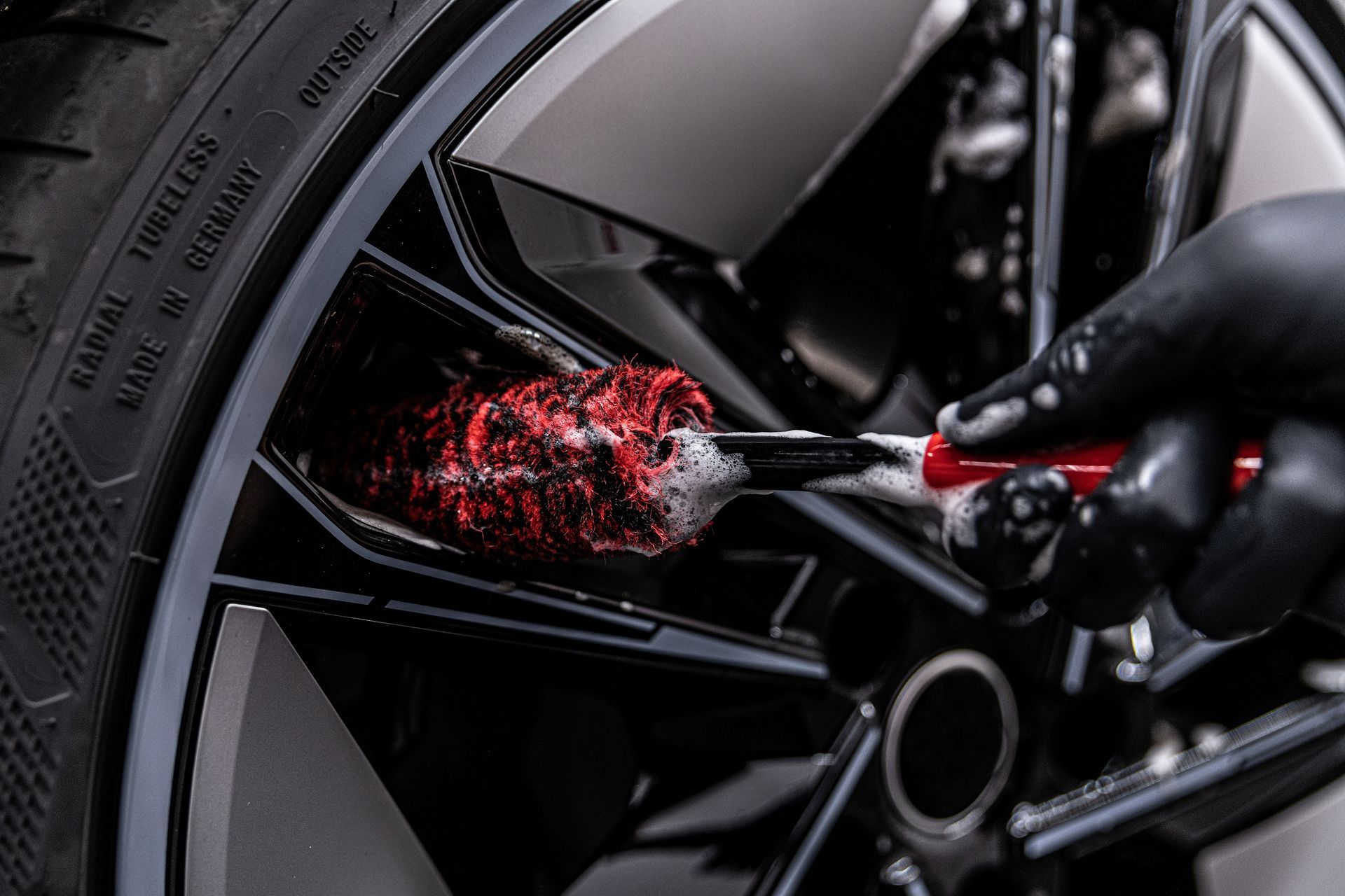 Black-gloved hand cleaning a car wheel with a red brush and soap suds.