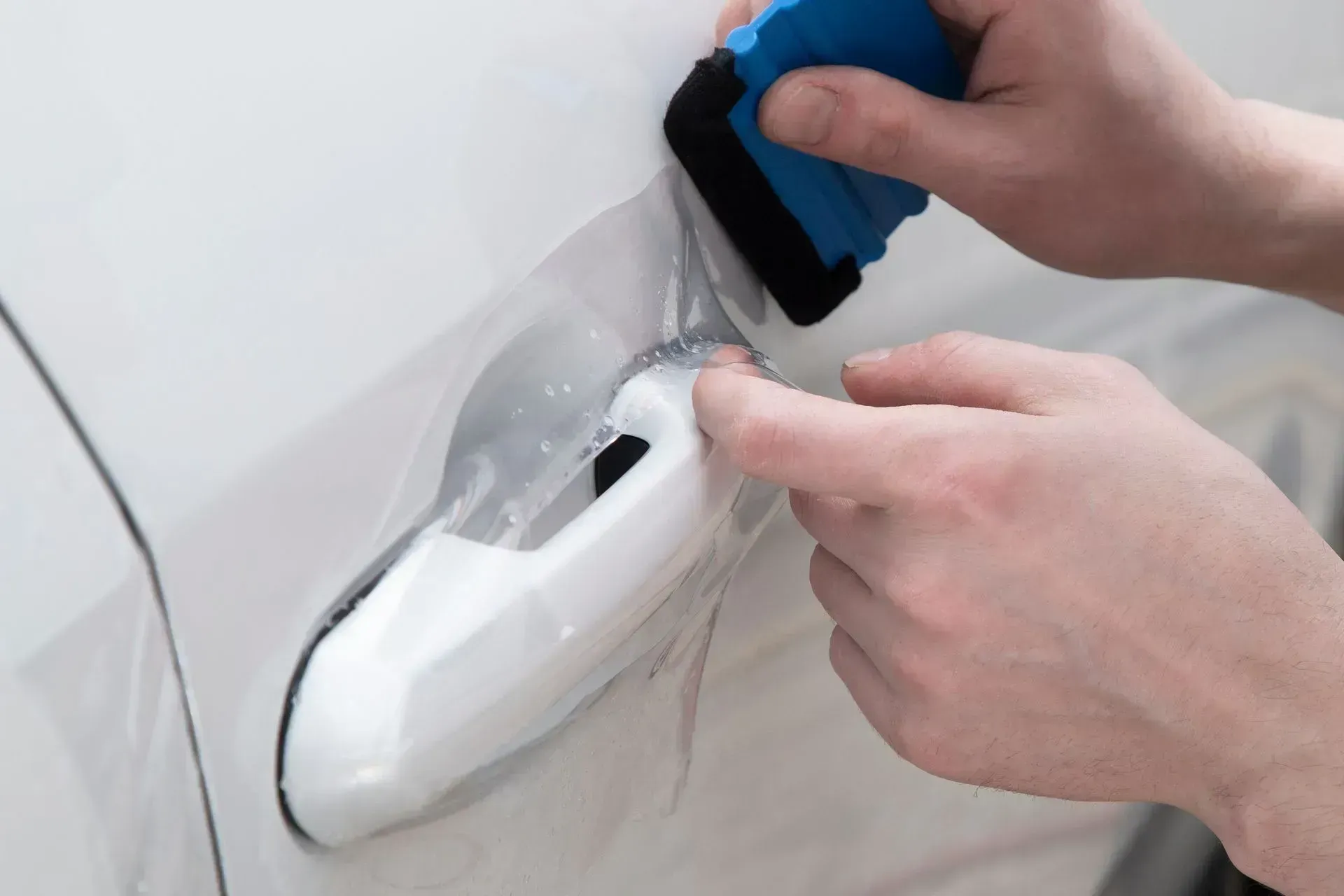 Person applying clear protective film to a car door handle with a blue squeegee.