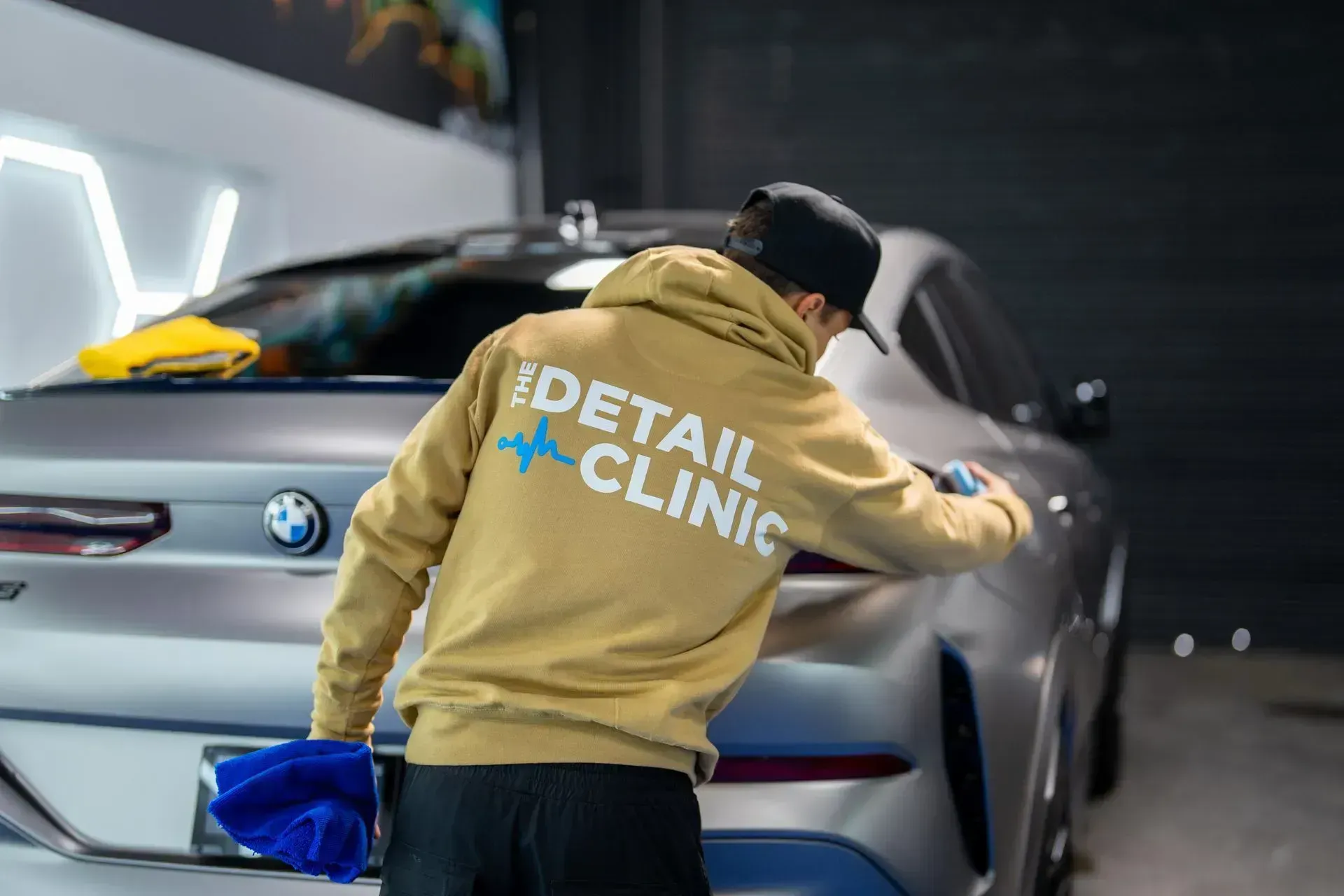 Person in tan hoodie cleaning a silver BMW at a detailing clinic.