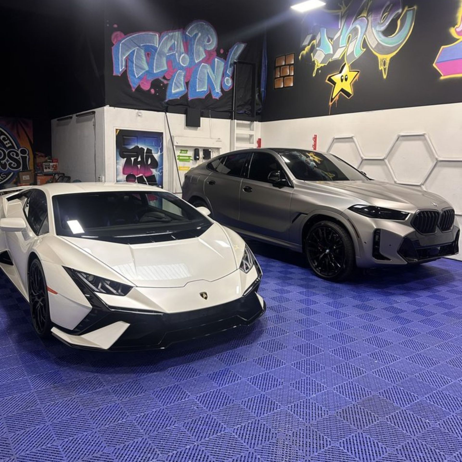 White Lamborghini and gray BMW inside a garage with graffiti art on the wall.