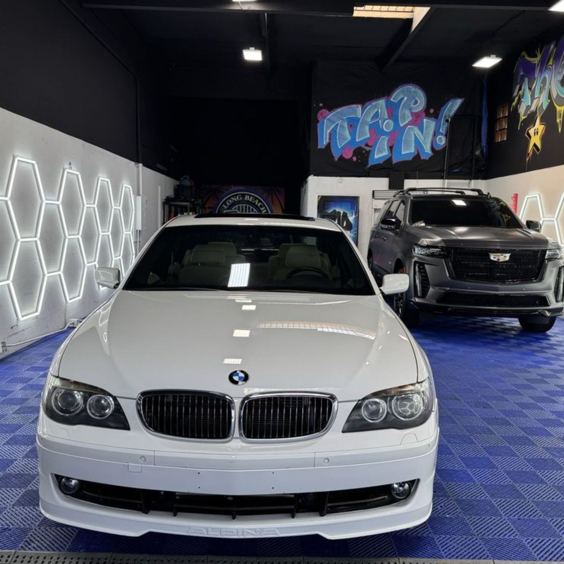 White BMW sedan in a detailing shop, black SUV in the background. Hexagon lights on wall.