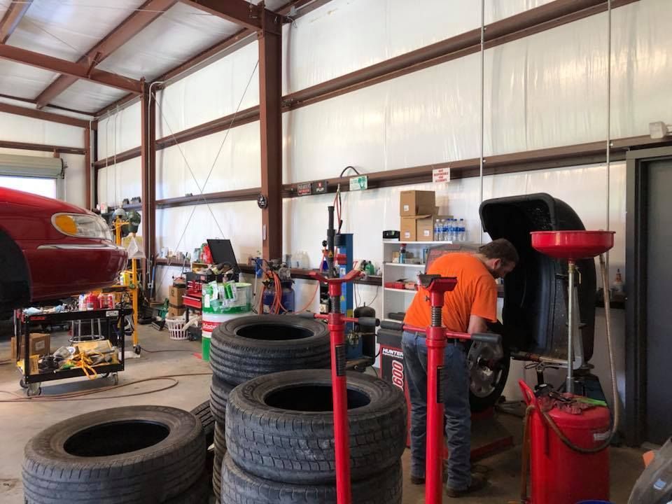 Mechanic working on a car in a shop. Tires stacked nearby, red car raised on lift, tools, and metal framework.