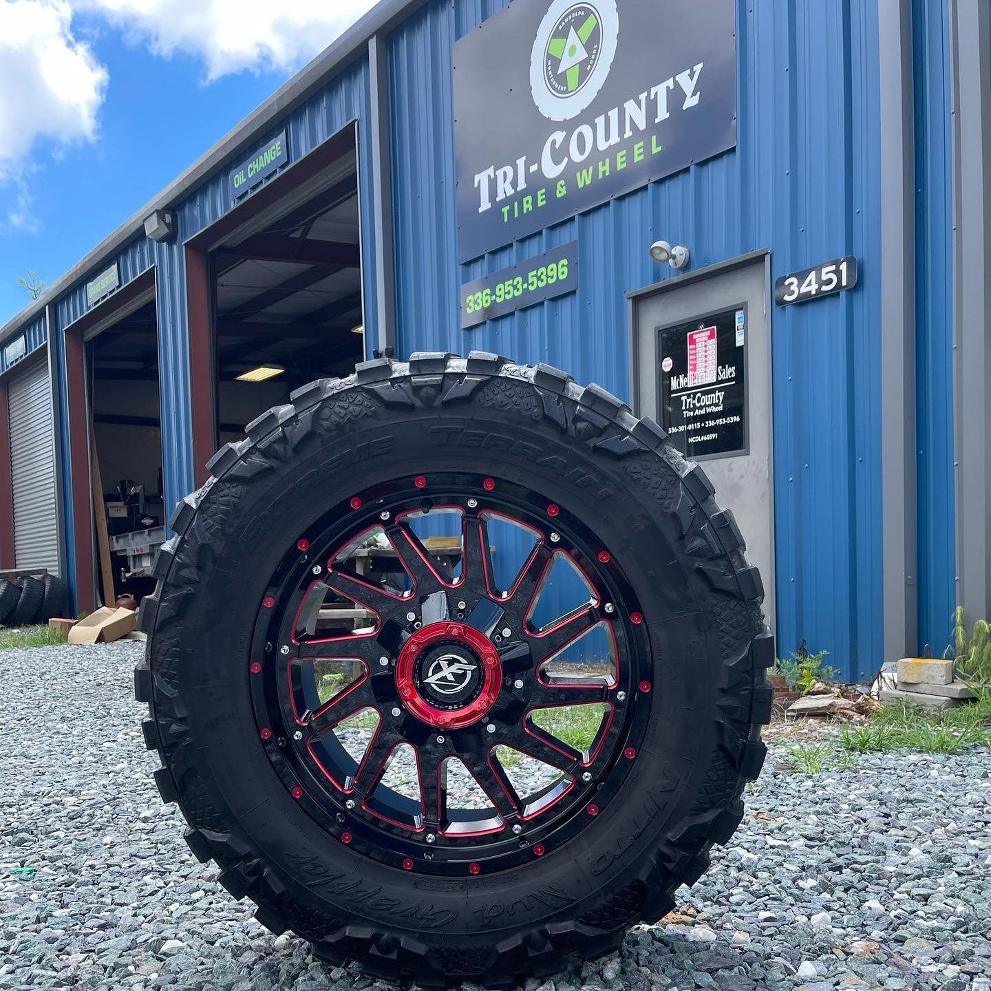 Large tire with a red and black rim in front of a blue Tri-County Tire & Wheel building.
