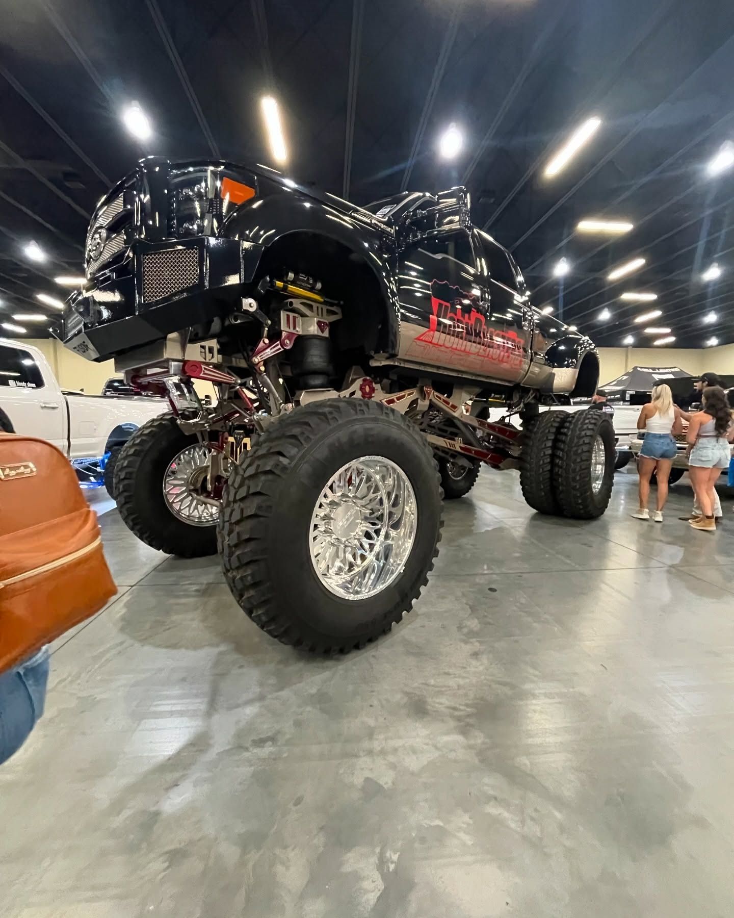 Monster truck with chrome wheels and black body at a show, people in background.