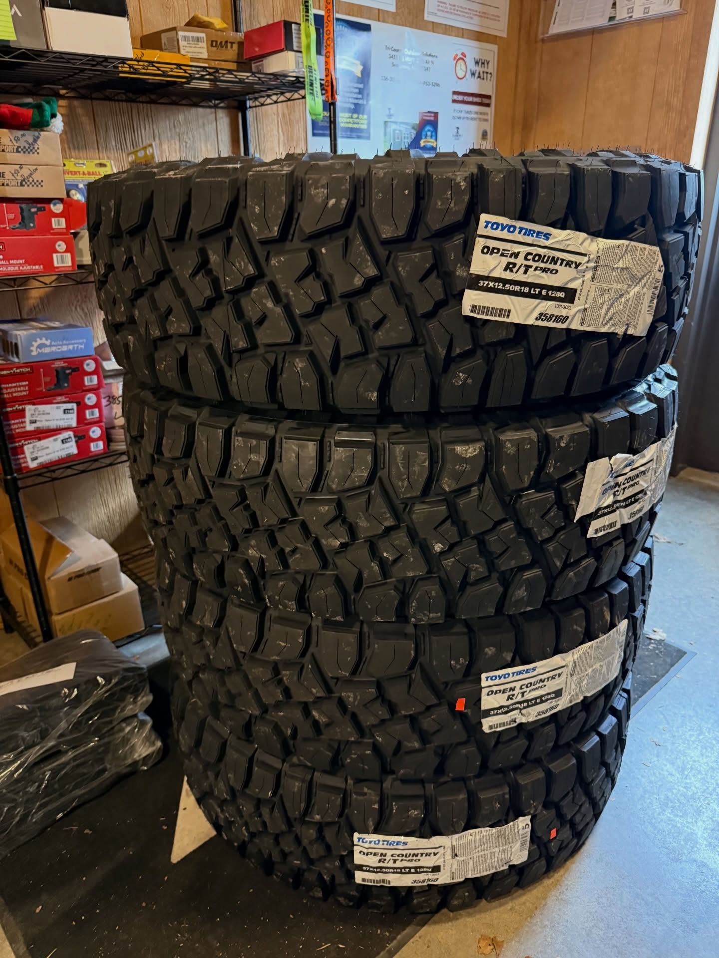 Four new black off-road tires stacked in a shop, with labels attached.